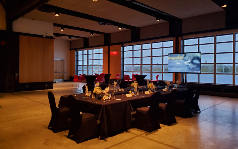 Banquet tables with chairs are placed in a U-shape facing a view of the harbor through windows. A tv is set-up for a presentation.