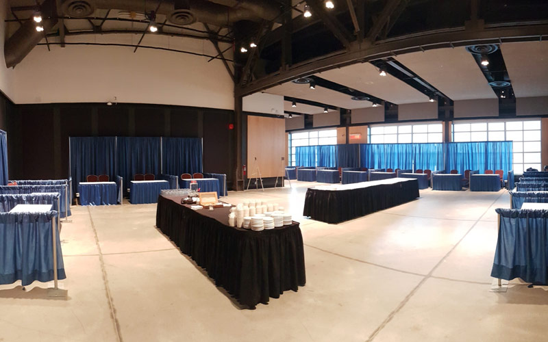 Tradeshow booths with blue skirted tables and two chairs each are divided by pipe and blue drapes. Two rectangular tables are set in centre of the room for beverage and snack stations.